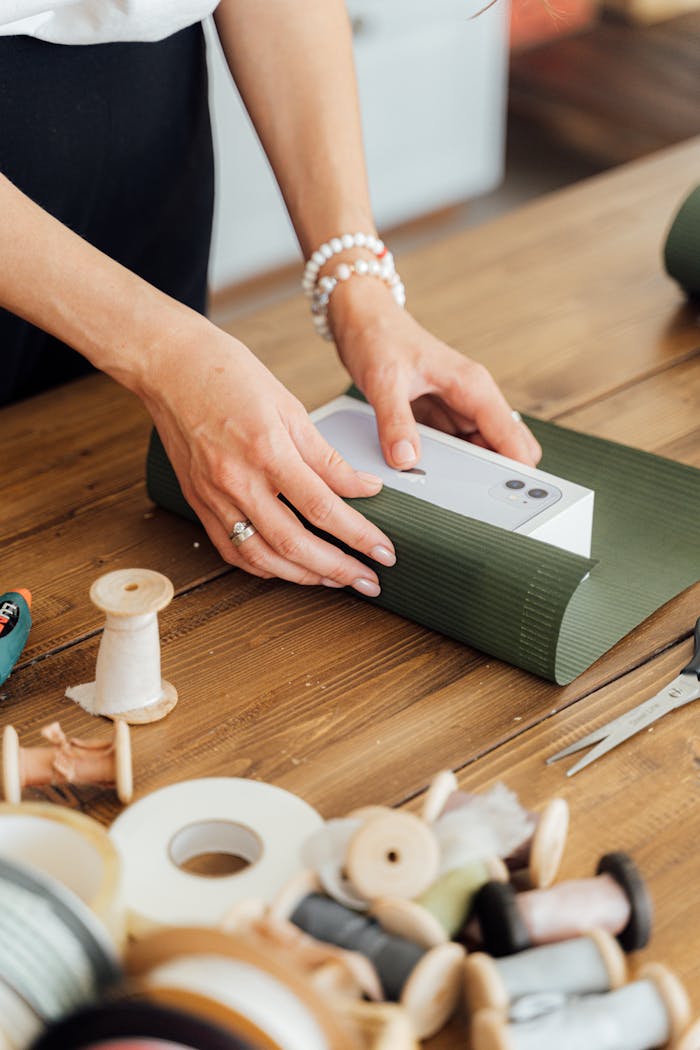 Close-up of hands wrapping a gift box with green paper on a wooden table, surrounded by sewing supplies.